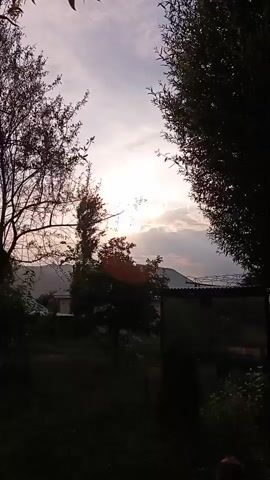 This is a short video clip showing a sunset. The sun is visible between clouds, casting a warm glow. Silhouettes of trees and hills frame the scene. The foreground is dark and blurred, with some foliage visible. The sky is a gradient of light colors, transitioning from yellow and orange near the sun to purple and grey further away.