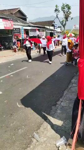 A group of people are marching in a procession on a paved road. Most of the participants are wearing white shirts and red headscarves. They are carrying a long, wide, red and white banner, which appears to be a flag. Some individuals in the front are holding the flag taut, while others behind them are also holding it. Several children are present, with one child in a red and white shirt walking alongside the adults holding the flag. The environment appears to be a village or small town, with buildings and shops lining the street. The sky is blue and the sun is shining, casting shadows on the road. There are spectators visible on the side of the road, some sitting and some standing. The scene suggests a celebratory or patriotic event.