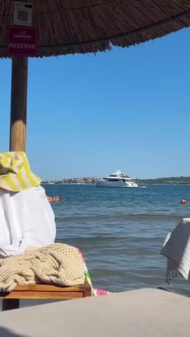 The video shows a beach scene from the perspective of someone lying on a lounge chair under a thatched umbrella. In the foreground, a beige knitted blanket and a white garment are draped over the lounge chair. A person wearing a yellow and green striped hat is partially visible in the background on the left. The water is calm and blue, with a white yacht sailing in the distance. A person is paddling on a paddleboard in the middle ground. A sign with the word "RESERVE" is attached to the umbrella pole. The sky is clear and blue.