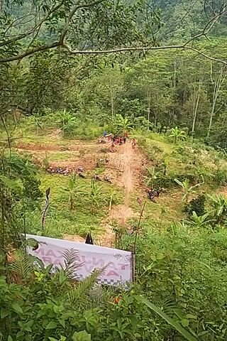 A camera pointed vertically downward films a dirt motorcycle race. A dirt course goes through the trees on a steep hillside. The cameraman is hidden in the brush at the side of the course, with spectators just to the left. A motorcycle and driver approach the camera, kicking up dirt. After the motorcycle passes, the camera moves to show the spectators.