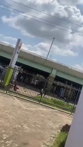 The video shows an outdoor scene with a cloudy sky. In the foreground, there's a black metal fence running parallel to the bottom of the frame. Behind the fence is a paved area with grass and some visible vegetation. In the middle ground, there's a tall, thin sign with red and white coloring, possibly a brand marker. Behind the sign is a light green overpass structure. Below the overpass, there's an area that appears to have several people present and some clothes hanging. There are some poles in the distance as well.