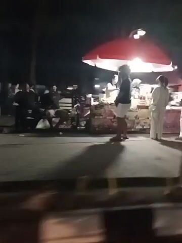 The video shows an outdoor night scene with several stalls and people. In the first frame, two people are standing at a stall with a red and white umbrella, illuminated by a bright light. Other people are visible in the background, some sitting and some standing. The second frame shows a person on a scooter near a stall. The third frame displays colorful lights from a stall. The fourth frame shows a person walking past a stall. The fifth frame shows people sitting on benches near a stall. The overall scene suggests a marketplace or a gathering at night.