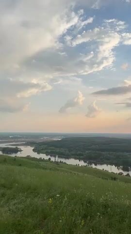 The video opens with a wide shot of a verdant hillside overlooking a wide river that winds through a dense forest. The sky is a mix of blue and white clouds, with hints of pink and orange suggesting either sunrise or sunset. The camera pans slowly to the right, revealing more of the river and the expansive landscape. As the camera continues to move, a section of a red brick ruin, resembling an archway, comes into view on the hilltop. The ruin is weathered and partially overgrown with grass. The camera stops to focus on the archway, and a small, brown animal, possibly a deer or a goat, is visible standing within the arch. The sky continues to display a dramatic interplay of light and shadow from the sun peeking through the clouds. The overall scene is serene and natural, with the ruin adding a touch of historical intrigue to the picturesque landscape.