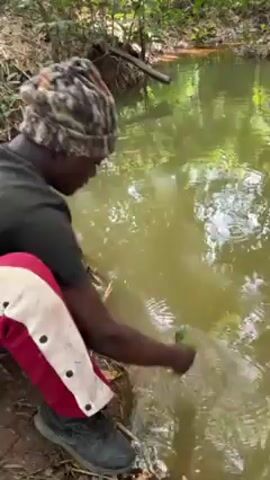 A person in a camouflage hat, black shirt, red pants with white stripes, and dark shoes is crouched by a narrow stream in a wooded area. They are using a green leaf to splash and stir the water near the stream's edge, creating ripples. The water is somewhat murky. The stream is surrounded by vegetation and fallen leaves. There is a fallen branch visible further along the stream.