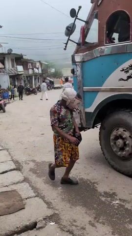 The video shows a person in a colorful outfit and a headscarf climbing onto the side of a large truck. The truck has a blue and white design on its side, including a dragon image. The person is holding something black in their left hand. The scene appears to be a street with other people and buildings in the background. The ground is wet and appears to be a dirt or gravel road. The weather seems overcast.