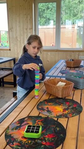 A young girl with long blonde hair pulled back in a ponytail, wearing a dark blue hoodie and light blue jeans, stands at a wooden table in a room with wood-paneled walls and large windows. She is focused on a tower of colorful plastic rings that she is building. The tower is approximately 15 rings high and is placed on the table in front of her. To her right, there is a woven basket containing white napkins. On the table are several round placemats with colorful bird and nature designs. In the foreground, there is a small green game with a grid pattern. The girl is in the process of adding another ring to the top of the tower, holding a single ring between her thumb and forefinger. The room appears to be an indoor recreational space, possibly a cafe or activity center, with other tables and chairs visible in the background. The lighting suggests it is daytime, with natural light coming through the windows.