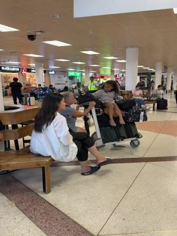 The video shows three women at an airport, one of whom is a child. The child sits on top of a luggage cart loaded with suitcases. A woman, presumably the child's grandmother, interacts with the child, holding her hand. Another woman sits on a nearby bench. The floor has a geometric tile pattern. Airport signage and personnel can be seen in the background.