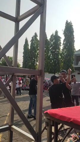 The video shows an outdoor scene with a metal structure in the foreground. In the background, a group of people are gathered. Some individuals are wearing black shirts and hats, and one person is holding up a phone, possibly recording or taking pictures. A group of women in red and white attire, some wearing headscarves, are performing some sort of synchronized movement, possibly a dance or exercise routine, in an open paved area. Trees and buildings are visible in the distance under a clear sky. A red and white ribbon is draped over the metal structure.