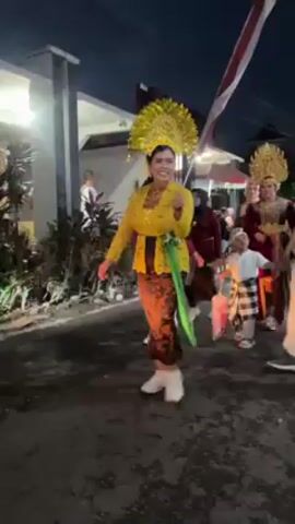 The video shows a woman dressed in traditional Indonesian clothing walking and smiling, apparently participating in a parade or festival. She wears a yellow long-sleeved top with lace details, a dark sarong-like skirt, white sneakers, and a bright yellow, halo-shaped headdress. A green sash is tied around her waist. She makes hand gestures toward the camera as she walks, appearing cheerful and engaged. Other people, some in similar traditional attire, are also present, walking alongside and behind her. In the background, there are houses, plants, a parked blue motorbike, and a red and white Indonesian flag. It appears to be dusk or early evening.