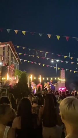 A crowd of people stand in front of a stage at night. On the stage, a band performs with musicians playing instruments and singers. The stage is lit with bright lights, and colorful flags are strung across the sky above the crowd and stage. A building is visible in the background to the left of the stage.