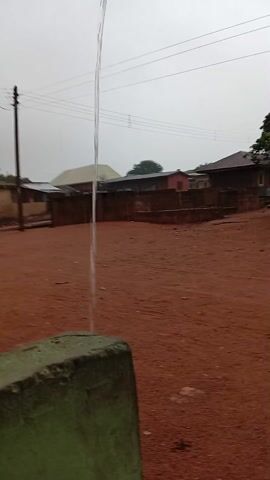 The video shows a view from a moving vehicle in a rural area. The ground is reddish-brown dirt. There are simple houses with brick walls and metal roofs, and trees. The sky is overcast and it looks like it might be raining. The camera initially focuses on a concrete post before moving. The view is partially obscured by the vehicle's frame.