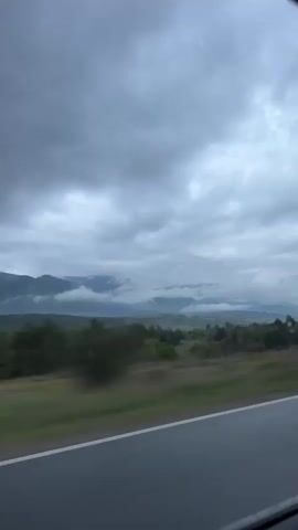 A roadside view from a moving vehicle is captured on an overcast day. The pavement and its white line markings are visible along the bottom of the frame. A metal guardrail runs along the side of the road. Beyond the guardrail, there is a stretch of green land with clusters of trees. Further into the distance, mountains are visible, partially obscured by low-hanging clouds. The sky is filled with dark, grey clouds. Another vehicle appears momentarily in the reflection of the car window.