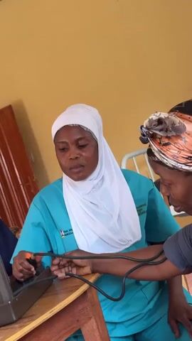 A woman in a teal scrub top and a white hijab is taking the blood pressure of another woman. The woman taking the blood pressure is sitting at a wooden table and holding the blood pressure cuff bulb. The other woman is standing next to the table and looking at the medical equipment. Behind them is a yellow wall and a hospital bed. The woman in scrubs appears focused on the procedure.