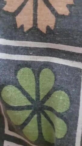 The video shows a close-up, slightly shaky, static shot of a fabric with a repeating pattern of stylized flowers. The fabric appears to be dark gray with a white border. Within the squares formed by the white border, there are two distinct floral designs. The top square contains a brown or tan flower with eight petals, each petal having a notched tip. The bottom square contains a green flower with eight rounded petals arranged in a circular pattern around a dark center. The fabric is slightly creased, indicating it is likely clothing or bedding. The lighting is even and diffused, with no harsh shadows. The video is very brief, lasting only a few seconds.