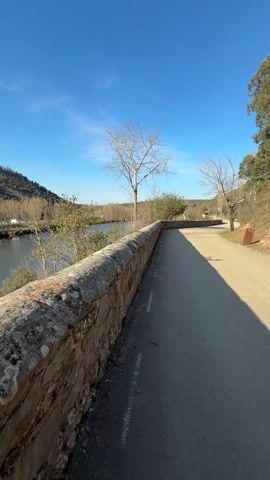 The camera moves from right to left, showing a road running alongside a stone wall. On the other side of the wall is a body of water reflecting the sun. The water is surrounded by gently sloping hills and sparse vegetation. The sky is clear blue.