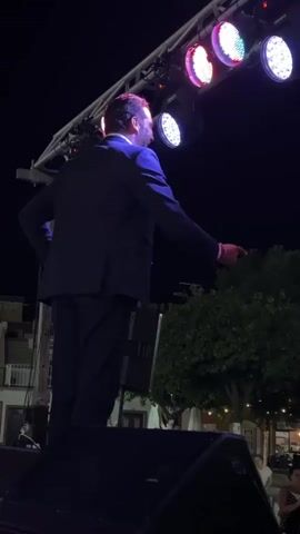 A man in a suit stands on a stage at night, facing away from the camera. He is illuminated by stage lights that are visible above and behind him. The man's right hand is slightly raised, and his left hand is also extended. In front of him is a large speaker. In the background, there are trees, buildings, and more lights. The sky is dark.