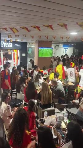 The video shows a crowded indoor space, likely a bar or restaurant, where a group of people are watching a soccer game on a television screen. Many individuals are wearing Spanish national team colors, with red and yellow flags draped around them and hanging from the ceiling. The atmosphere appears energetic and excited, with people clapping and cheering. Some patrons are seated at tables, while others are standing. The room is illuminated by overhead lights and the glow from the television. The overall impression is one of a shared, enthusiastic viewing experience of a sporting event.