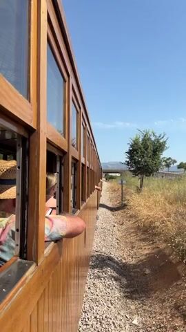 A person wearing a straw hat leans out of a train window, their arm resting on the windowsill. The train is made of polished wood and has large windows. Another person, wearing a brightly colored shirt, is also visible inside the train, leaning out of the window and touching the side of the train car. The train is moving along a track bordered by gravel and dry vegetation. In the distance, there is a bridge, some trees, and mountains under a clear blue sky. The camera is positioned to show the side of the train from a low angle, looking forward along the track.