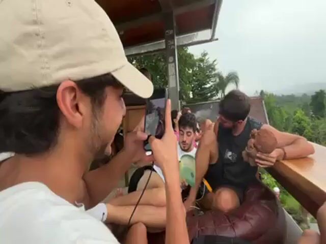 The video shows a group of young people on a covered patio. In the foreground, one man is holding a smartphone and appears to be taking a photo of another man who is lying on a large brown cushion reading a book. A third man is seated next to him at a table, holding a coconut. The man with the phone is smiling and the man reading is smiling at the phone. Other people are sitting at tables in the background. The patio has a view of a green, forested landscape in the distance. The weather appears overcast.