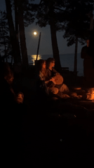 Nighttime campfire gathering by the water, moon overhead