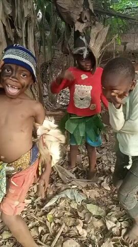 The video shows three children in an outdoor setting with dry leaves and banana trees. One child is partially unclothed, with dark face paint and a striped hat, wearing a patterned wrap around the waist, and holding something white. Another child, also with dark face paint, wears a red shirt, and a leaf skirt. The third child wears a light-colored long-sleeved shirt and pants. All three children are barefoot and appear to be posing or playing for the camera. The child in the long-sleeved shirt covers their face with their hands. The backdrop includes greenery and some visible structures.