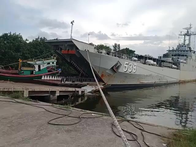 The video shows a naval vessel, hull number 539, docked at a concrete pier alongside a smaller wooden fishing boat. A ramp extends from the vessel onto the pier. The camera pans to the right, revealing a large, blue and white ferry named "Wira Loewisa" also docked at a nearby pier.  In the background are industrial structures including warehouses, silos, and a crane against a cloudy sky. The water in the harbor is relatively calm. There is vegetation along the edge of the pier in the foreground.
