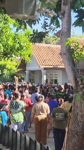 A crowd of people, including many women and children, is gathered in an outdoor setting with a house visible in the background. They are reaching their hands up in the air, appearing to be trying to catch something. The camera is positioned behind a fence and partially obscured by trees and hanging plants. The house is single-story with a tiled roof and white pillars. The setting appears to be a residential area on a sunny day.