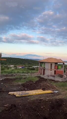 The scene captures a construction site featuring partially-built structures against a backdrop of a vibrant sky during the late evening. 

In the foreground, there are mounds of dark soil and scattered construction materials, including yellow wooden planks, on the ground. 

The central focus is a two-story building under construction. Its structure is partially completed, with concrete blocks and some brickwork visible. There's a rudimentary wooden roof framework already in place. The building’s design includes what appears to be a balcony or overhanging structure.

Further back, other foundations and concrete structures are visible, indicating that multiple buildings are being constructed on the site. There are also wooden structures set up as scaffolding.

In the distance, the landscape reveals a mix of green fields, sparse vegetation, and a body of water, possibly a lake or river, with a gentle shoreline. The sky above is a blend of colors, featuring shades of blue, white, and pink, suggesting either sunrise or sunset. There are no people visible in the frame.