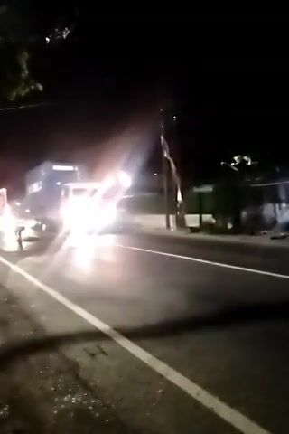 A nighttime video shows a truck with a blue shipping container on a road. The truck's headlights are bright, and the container has writing on it that is partially visible. There are other lights in the background, suggesting other vehicles or streetlights. The road appears to be paved, and there is foliage visible on the side of the road. The video is shaky, suggesting it was recorded by someone moving.
