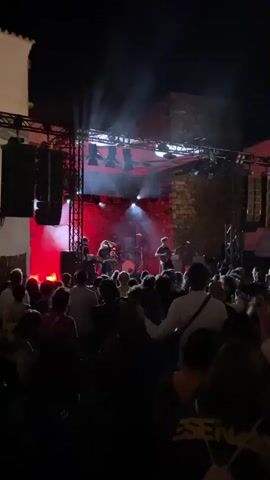 A live band performs on an outdoor stage at night, illuminated by green and red stage lights. A crowd of people stands in front of the stage, some holding up phones. The stage is set up with musical instruments, including drums and guitars. The background shows stone walls and a dark sky. The video captures the energy of the concert, with the musicians actively playing and the audience appearing engaged.