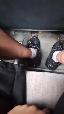 The video clip begins with a close-up shot of a person's feet, likely a bus driver, wearing black sneakers and white socks. One foot is positioned near what appears to be accelerator or brake pedals, resting on a worn, somewhat dirty floor with visible metal parts in the footwell.

The camera quickly pans up to reveal the interior of a passenger bus, busy with people. The scene is set in Kornitsa, Bulgaria. The bus is well-occupied, with numerous passengers, including adults and possibly younger individuals, either seated or standing. The seats are upholstered in a purple and grey fabric with an abstract, colorful pattern. Many passengers are visible from behind, looking forward, while some are standing in the aisle, holding onto overhead handrails as the bus appears to be in motion. Through the windows, green trees and outdoor scenery suggest travel through a rural or suburban area. The time of day is daytime, with good natural light. Passengers appear calm and engaged in their journey. A young male passenger in a plaid shirt is visible, as is another person in a blue hoodie standing in the aisle. No discernible text is visible within the bus interior or outside.