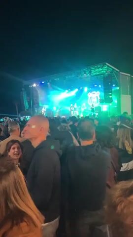 This video shows a live outdoor concert at night. A band is performing on a raised stage with green and blue lighting illuminating the stage and the band. The band's logo is displayed above them. A large crowd has gathered to watch the performance, with many people facing the stage. The crowd appears dense, with individuals standing closely together. Tents and flags with logos are visible at the back of the crowd. The overall atmosphere is vibrant, with a focus on the live music and the audience's experience.