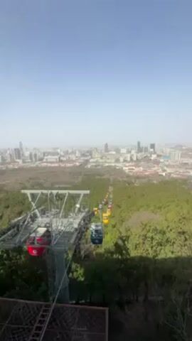 The video shows an aerial view of a city and a forest from a cable car station. In the foreground is a cable car station with a tall metal tower and red and blue cable cars. The cable cars are attached to a cable that stretches into the distance. The forest is below the cable cars and is dense with green trees. In the background is a city with many tall buildings. The sky is blue and clear.