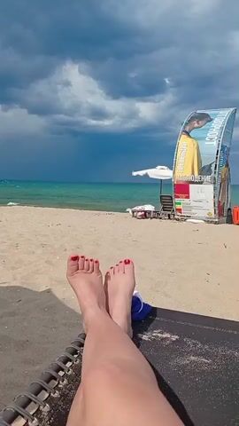 A first-person perspective video shows a person's feet resting on a beach lounge chair. The person's toes are painted red. The background features a sandy beach with two white beach umbrellas, lounge chairs, and a dark, cloudy sky over the turquoise ocean. In the distance, several kitesurfers are visible on the water. A large, colorful kiteboarding banner is to the right of the frame.
