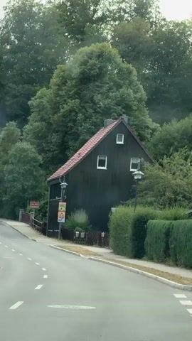 A drone view shows a dark wooden house with a red roof nestled amongst lush green trees. A road curves past the house. The house has three windows on its front facade, two on the upper level and one on the lower level. There are also two light fixtures attached to the exterior of the house. A signpost with a red sign is visible to the left of the house, and two colorful banners hang from a pole in front of the house. The road is paved and has white dashed lines marking the lanes.