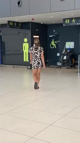 A woman in a black and white patterned romper walks away from the camera towards a dark wall with accessible restroom signs. To her left is a restroom sign with a male figure in a wheelchair. To her right is a janitorial cart with cleaning supplies and trash bags. She walks past the cart and reaches for something on a metal partition. She then continues walking away from the camera.