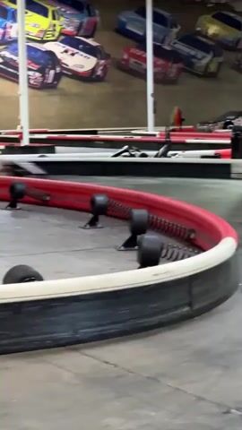 The video shows a go-kart track indoors. Two go-karts, one primarily black and another with a lighter color scheme, are actively racing on the track. The racers are wearing helmets. The track is bordered with red and white barriers, with black rollers attached to the barriers. The indoor space is large with a high ceiling, supported by metal beams. Wall decorations depicting race cars are visible. Signage and a large display screen are also present in the background. The camera follows the racers as they navigate a curve in the track.