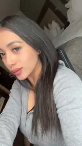 The video shows a young woman with long, straight, dark hair, wearing a gray zippered sweatshirt. She is sitting in a chair, and a bed is visible in the background. She is looking at the camera and moving her head slightly.