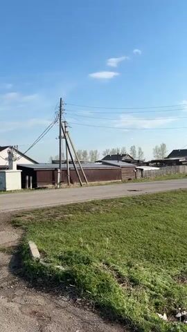 The video shows a rural landscape under a bright blue sky with scattered white clouds. A paved road curves gently uphill, bordered by green grassy verges. Utility poles line the road, carrying wires overhead. On either side of the road, there are houses and outbuildings typical of a village setting. The buildings vary in appearance, some made of wood, others with metal siding. The scene is static, with no visible movement or activity.