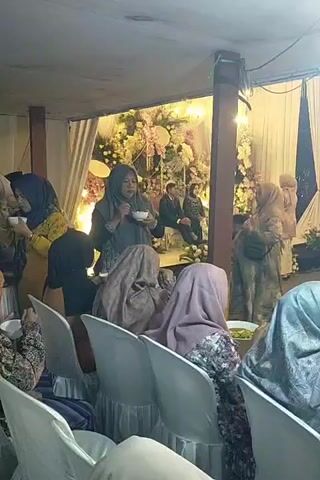 A group of women are seated in white chairs facing a decorated backdrop. In the center of the frame, a woman wearing a grey hijab is holding a small bowl and spoon, appearing to eat. To her right, another woman in a light pink hijab is seated. Behind them, a man and two women are seated on chairs near the backdrop, which is adorned with flowers and fabric. To the far right, a woman in a beige outfit and hijab stands. The environment appears to be an indoor event space with draped fabric and lighting. The camera angle is from behind the seated women, looking towards the backdrop.