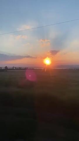 A time-lapse video shows a sunset over a rural landscape. The sun is visible in the center of the frame, casting a warm orange glow. Clouds are scattered across the sky, and the sun's rays are peeking through them. In the foreground, trees are blurred due to motion, indicating the camera is moving. The landscape appears to be a field with some scattered vegetation. The overall impression is a serene and beautiful natural scene captured during the transition from day to night.
