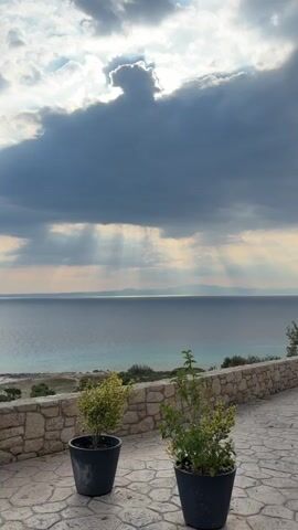 The video shows a view of the sea from a patio with two potted plants in the foreground. The sky is cloudy with sunbeams breaking through. The water is a deep blue and there is land visible in the distance. The patio is paved with stones and there is a stone wall along the edge. The potted plants are green and healthy. There is a black lantern hanging in the top left corner. There are a few birds flying in the sky. The scene appears to be tranquil and serene.