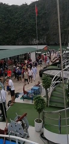 The video depicts a busy waterfront scene, seemingly in Vietnam, given the flag. A large group of people are walking along a pier with a green-roofed structure overhead. Many are wearing life vests. The scene is shot from the deck of a boat, looking onto the pier. There are chairs, railings, and potted plants on the deck, and a green spiral staircase leads to another level. In the background, a mountain covered with green vegetation rises from the water. The overall impression is a bustling scene of tourists getting on or off boats.