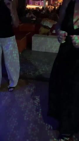 The video begins with a close-up shot of a person in a black dress, with flashing pink and green lights projected onto them. The background is blurred, but appears to be an indoor environment with furniture. The scene then transitions to a medium shot of a woman with blonde hair wearing glasses and a black dress, smiling and looking at the camera. She is in an indoor setting with other people visible in the background, and there are red and white chairs. The video then cuts to a woman with blonde hair wearing a black sleeveless top and white pants, dancing and looking at the camera. The background shows more seating and people. Finally, the video shows a woman with short gray hair wearing a red polka dot dress and a denim jacket, making a peace sign with her fingers and smiling at the camera. There are people in the background, and the lighting appears to be blue. The overall environment suggests a bar, club, or party setting.