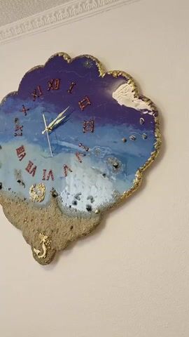 A decorative clock is displayed on a light-colored wall. The clock is irregularly shaped, resembling a cloud or a crest, with a scalloped edge. The clock face is designed to look like a beach scene, with a sandy area at the bottom and a blue ocean transitioning into a purple sky at the top. Roman numerals, in red, mark the hours. The clock hands, one long and one short, are gold. A small gold seahorse embellishment is attached to the sandy portion of the clock. The clock appears to be made of resin, with some glitter and small stones embedded in the design.
