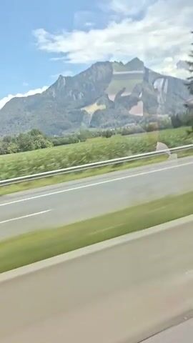 The video shows a view from a vehicle traveling along a road. The road has multiple lanes separated by white lines and a guardrail. On the left side of the road, there is a green field and a large mountain range in the background with scattered clouds. A few trees are visible in the foreground. On the right side of the road, a white RV is driving in the same direction. The sky is partly cloudy. The footage appears to be taken from a car or bus window.