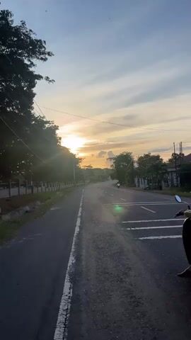 The video shows a person riding a scooter down a paved road. The sun is setting in the background, casting a warm glow across the sky and trees. The road is lined with trees on the left side, and buildings on the right. There are power lines running overhead. The scooter is silver and the rider is wearing a headscarf. The scooter is moving away from the camera.