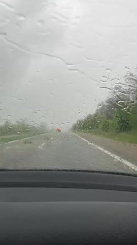 The view from inside a car shows a road with a red car ahead, during heavy rain. The road has a dashed white line in the middle. On either side of the road is some foliage and trees. The windshield has a lot of water drops on it, obscuring the view somewhat.