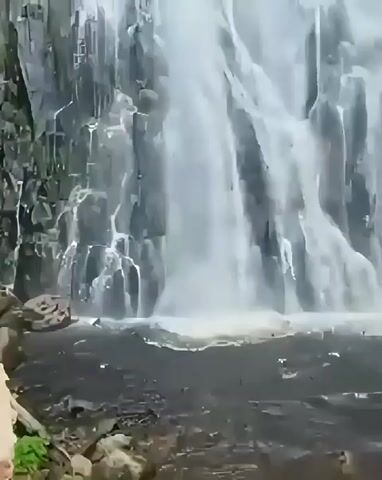 The video displays a stunning natural scene of a powerful waterfall cascading down a rocky cliff face. The water is white and frothy, creating a dynamic and energetic display. The surrounding environment is lush and green, with trees and foliage visible at the top of the waterfall, suggesting a tropical or subtropical setting. The rocky cliffs are dark and textured, providing a stark contrast to the white water. The lighting appears to be natural daylight, possibly overcast given the diffused light. There is no visible text in the video. The overall impression is one of natural beauty and raw power. The location is identified as Sosor Dolok, Indonesia.