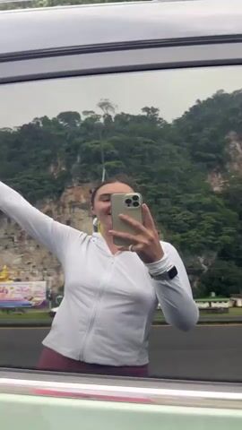 The video captures a young woman taking a selfie in the reflection of a car window. The woman is wearing a white zip-up jacket. She is smiling and raising her right arm in a fist-pump gesture. In the reflection, a lush green mountain with rocky outcrops serves as the backdrop. A road with a few cars is visible in the distance, along with some signs and buildings. The second part of the video shows the woman exiting the car, wearing a white crop top and bright pink pants. She is holding a purse and a red bag. The environment appears to be outdoors, with trees and foliage surrounding the area.